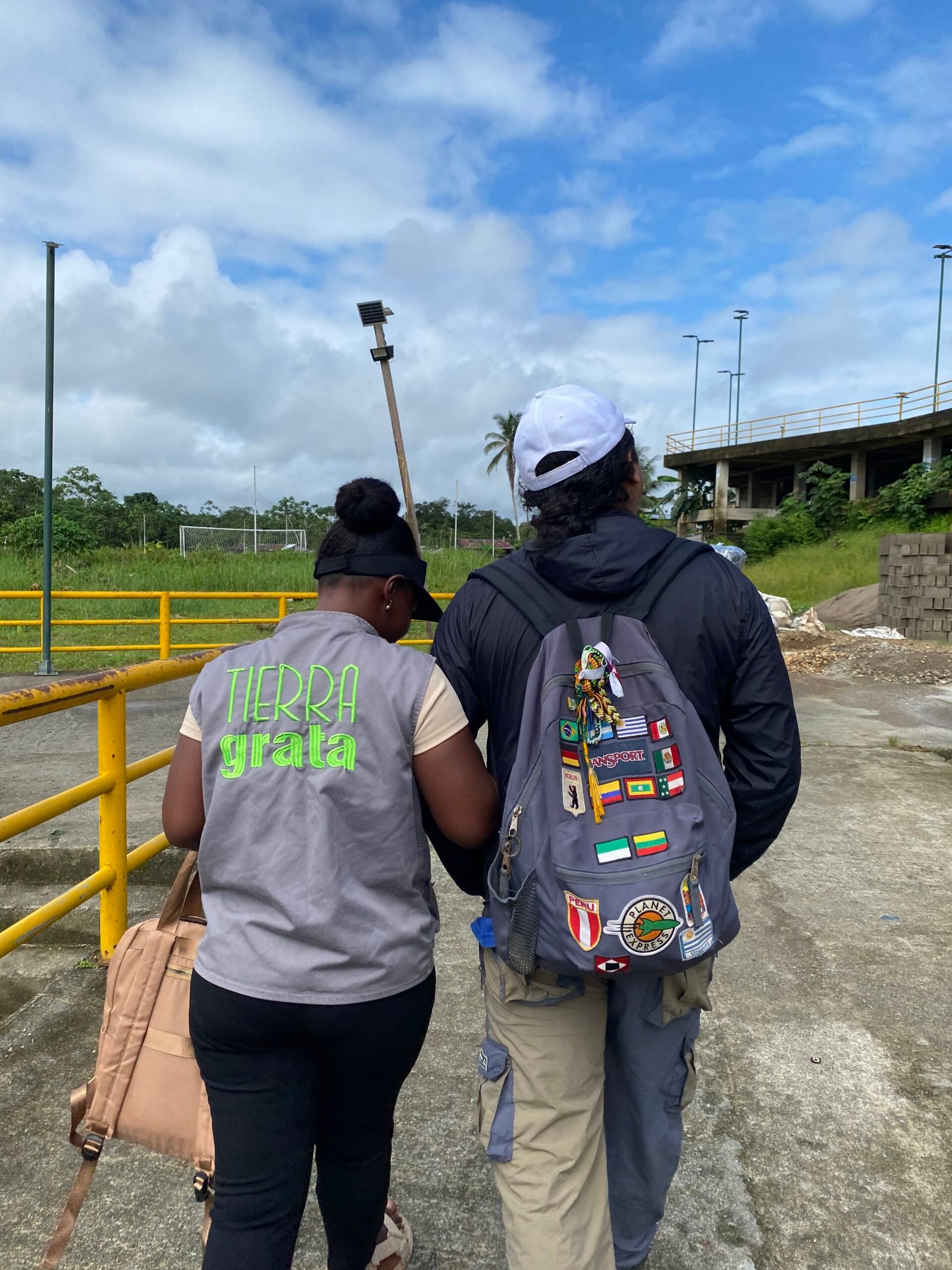 Dos personas caminan juntas por un espacio abierto en el Chocó. Una de ellas lleva un chaleco gris con el logo verde de Tierra Grata y un bolso beige; la otra viste una chaqueta negra, gorra blanca y una mochila con banderas de diferentes países. Al fondo se observan barandas amarillas, una cancha de fútbol, vegetación y un cielo parcialmente nublado.