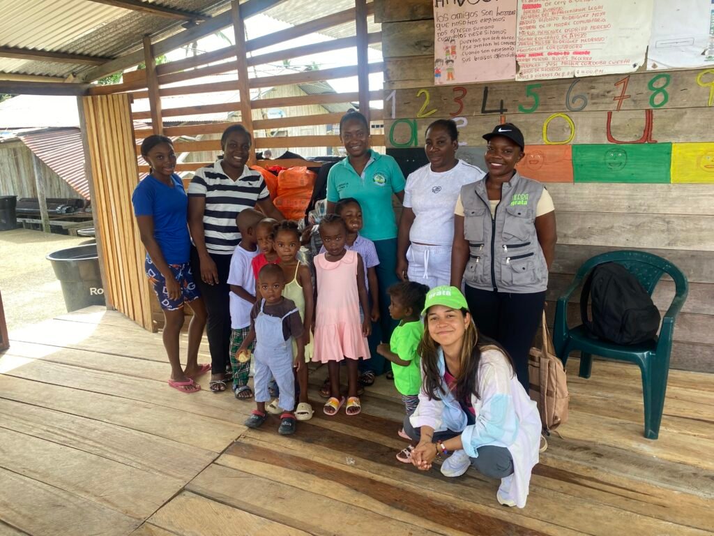 Fotografía grupal de integrantes de una comunidad rural del Chocó, junto a representantes de la Fundación Tierra Grata y organizaciones aliadas, durante una jornada de entrega de ayudas. En la imagen aparecen mujeres, niños y niñas en un espacio comunitario de madera, con materiales educativos en las paredes.